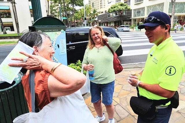 A man in a neon yellow uniform speaks to two tourists about directions.